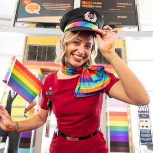 An air host waves a rainbow flag in front of a rainbow entrance gate (print, supply and installation by Ziggi's Print & Signs) for the Pride Flight to Mardi Gras in 2024.